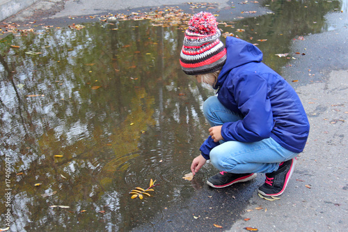 Fototapeta Naklejka Na Ścianę i Meble -  Kid girl in a warm knitted cap playing with leaves near a puddle while walking in an autumn park. Child wears a bright warm clothing on a cloudy autumn day.
