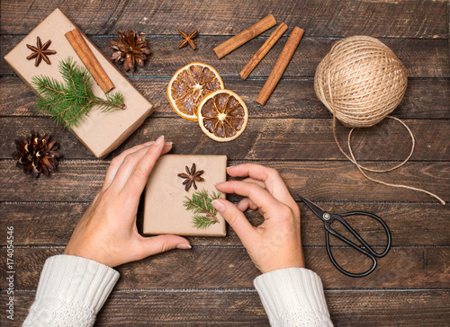 Woman decorating Christmas gifts. Presents wrapping inspirations. Hands, gift boxes, ball of jute, cinnamon sticks, anise, orange slices, fer tree branches and scissors on wooden background.