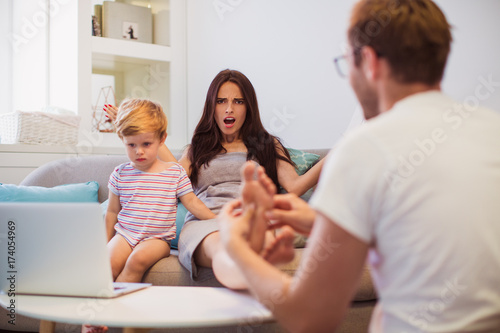 The young man doing foot massage his angry wife who sitting on the sofa near little son who watching laptop