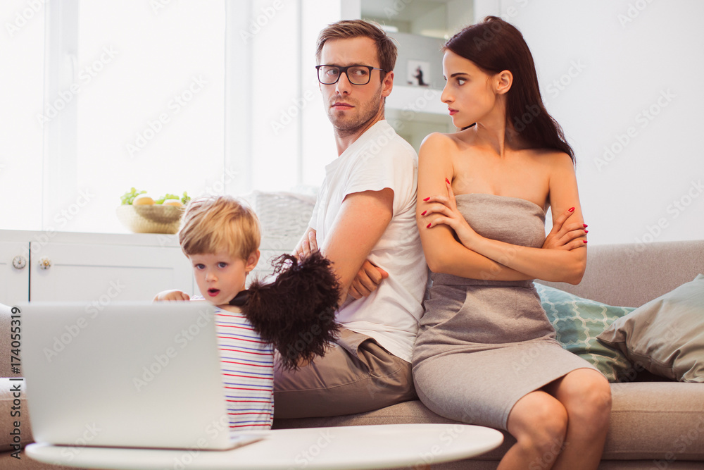 The young serious parents sitting on the sofa near their little son who watching laptop in the room The young serious parents sitting on the sofa near their little son who watching laptop in the room