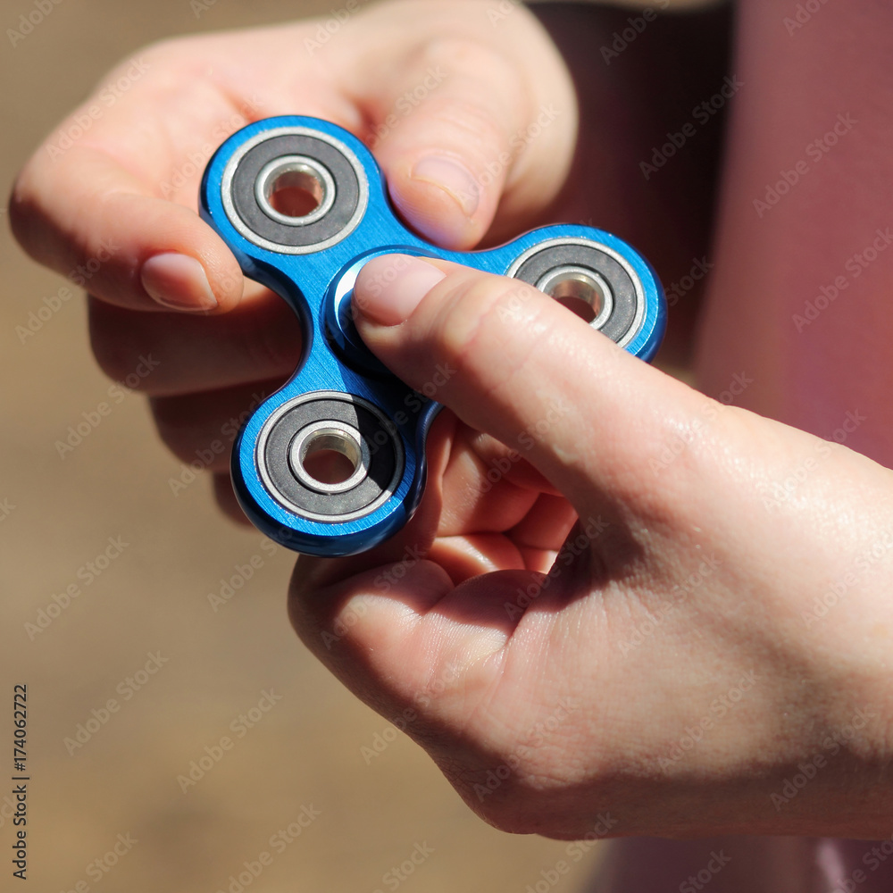 Fototapeta premium girl playing blue metal spinner in hands on the street, female hands holding popular fidget spinner toy on gray background, anxiety relief toy, anti stress and relaxation fidgets.