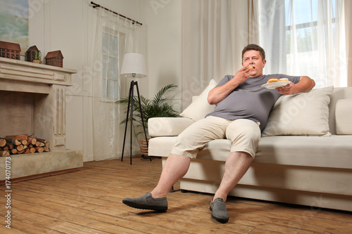 Photos Overweight young man eating sweets on sofa at home