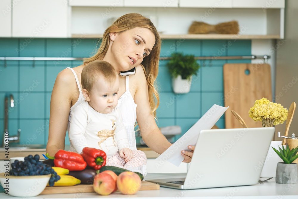Young mother is working home. Cooking and playing with her baby daughter in a modern kitchen setting. Healthy food concept. Looking at laptop.