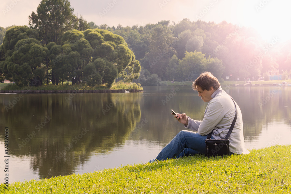 Man is sitting on the beach at lake and writing a sms on phone.