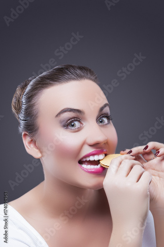Food Concepts. Portrait of Surprised Caucasian Brunette Female Biting Round Cookie. Demonstrating Surprised Facial Expression.Against Gray