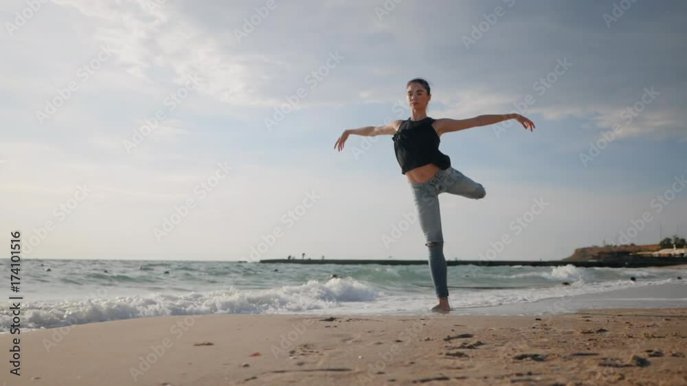 Young woman in casual style - denim and black top doing ballet at the beach. Attractive ballerina practices in stretching on sandy coastline in autumn. Slow motion. Young woman in casual style - denim and black top doing ballet at the beach. Attractive ballerina practices in stretching on sandy coastline in autumn. Slow motion.