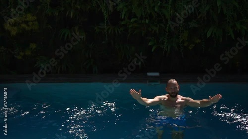Man standing in the swimming pool and splashing water, slow motion shot at 240fps, steadycam shot 
