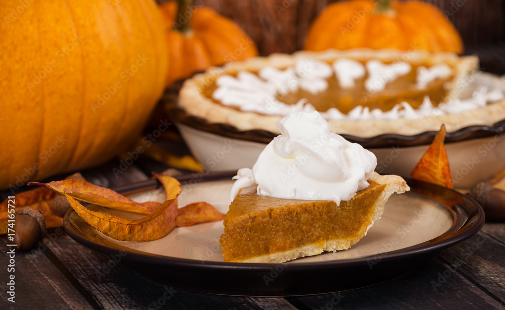 Slice of a pumpkin pie served with whipped cream. Pumpkins and autumn decorations displayed on rustic table. Slice of a pumpkin pie served with whipped cream. Pumpkins and autumn decorations displayed on rustic table.