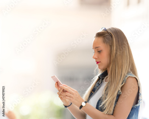 Pretty blonde teenager texting with her mobile phone outdoors in the city  defocused copy space background