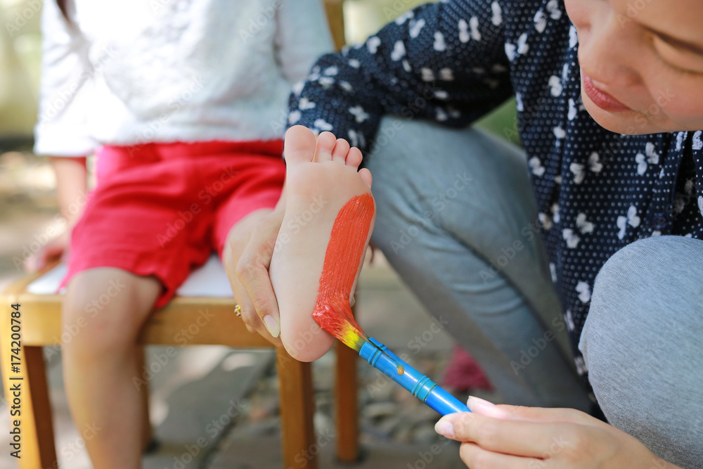 Fototapeta premium Mother painted foot with color green-orange for her daughter in the park. Focus at child foot.