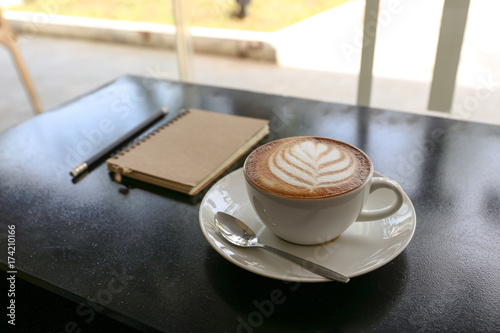 Hot latte coffee with leaf shape in white cup  notebook and pencil on the black metal table