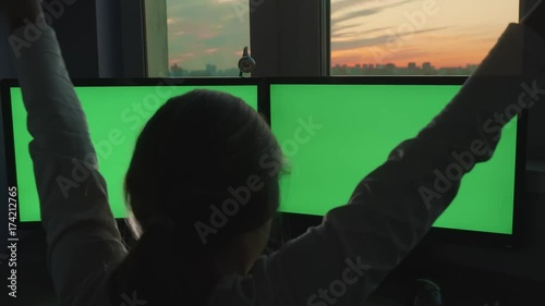 Blonde girl teenager throwing his hands up in front of large green screen monitors on backdrop of pink sunsets