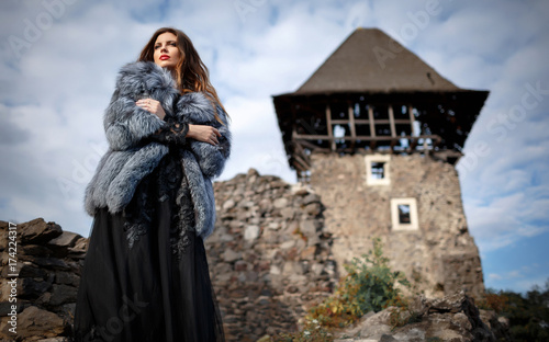 elegant brunette in a long black dress and coat stands on the background of a medieval castle