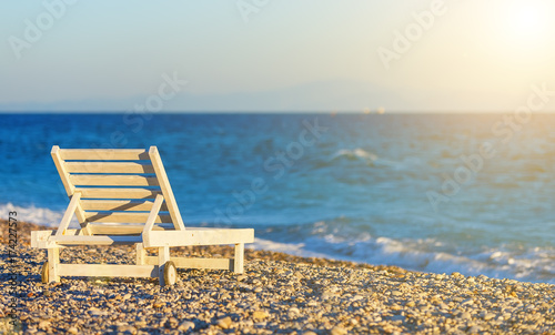 white wooden chaise longue on the shore of pebble beach at sunset, blur, depth of field © vladimircaribb
