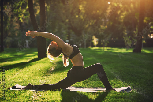 Beautiful young woman practices yoga asana Virabhadrasana 1 - warrior pose 1 in the park at sunset photo