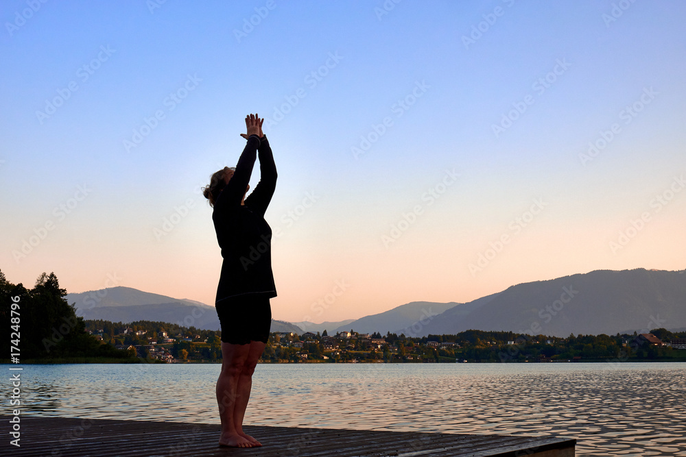 Fototapeta premium Mature woman in black clothes doing yoga on a wood jetty on Faaker See in Austria, early in the morning at sunrise