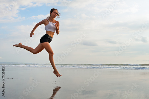 Barefoot sporty girl with slim body running along sea surf by water pool to keep fit and health. Beach background with blue sky. Woman fitness, jogging sports activity on summer family vacation.