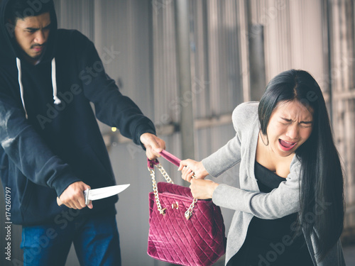 Culprit on hood holding knife is stealing bag from asian woman while walking alone at street, danger, burglar, criminal and robbery for woman concept.