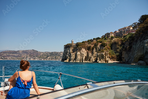 Boating, woman on deck in foreground, coastline bkgd.