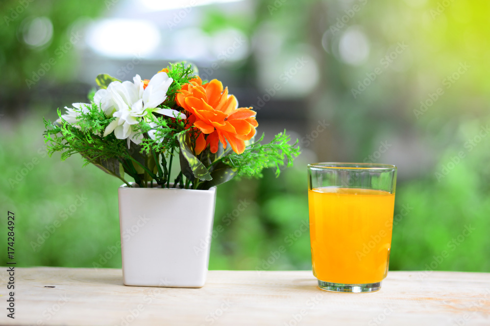 the white and orange  flowers in a white vase with a glass of orange juice on wooden table and yellow light in morning