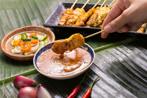 Schilderij op canvas A hand having pork satay dipping peanut sauce decorated with cucumber sauce, onion, chili on banana leaf