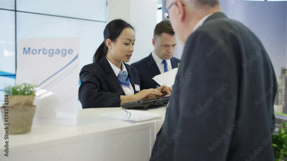 Bank worker at service desk working on computer & assisting customers Bank worker at service desk working on computer & assisting customers
