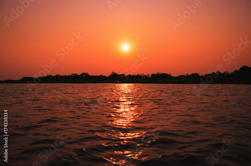 Amazing sunset at Paraguai river in Pantanal, Brazil. The river, the warm sun at sunset on a clear sky, water reflection of the light of sun and silhouette of trees on background.