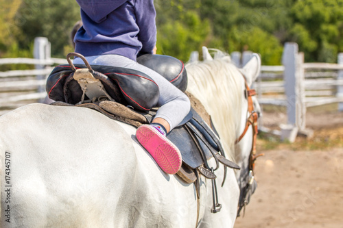 Little girl sitting in a saddle on a horse back and having fun riding along wooden fence at farm or ranch