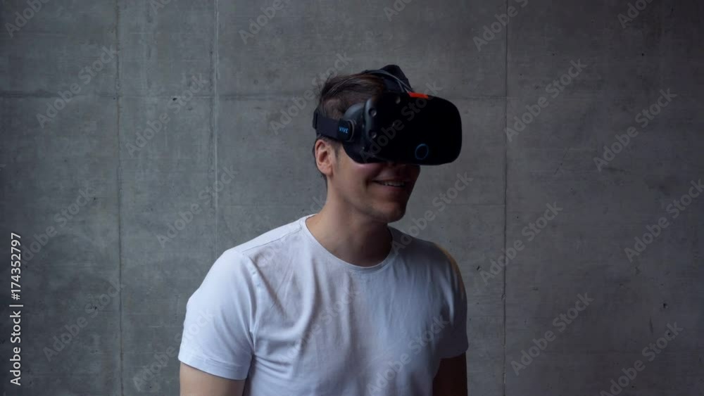 An adult male uses a virtual reality headset, standing behind a grey concrete wall. Medium shot. An adult male uses a virtual reality headset, standing behind a grey concrete wall. Medium shot.