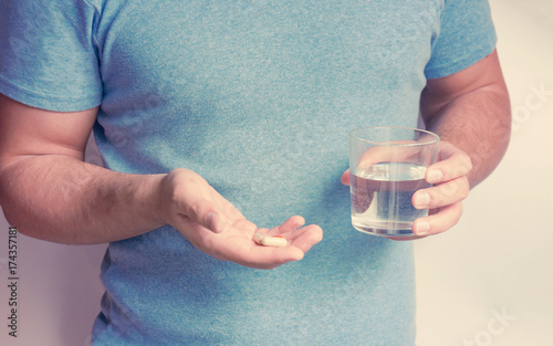 A young man in a blue t-shorts holds a group of capsules with medicine or vitamins in his hands and a glass of water in the other hand