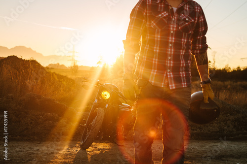 wild, young and free - deatail of a biker with helmet in his hand walking along a road at sunset with his bike in the back photo