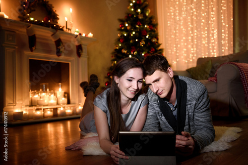 A cozy evening of a young family watching something together on a tablet, christmas decorations around in their room. Caucasian Couple using digital tablet at home celebrating New Year.