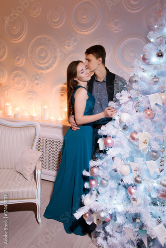 A tall handsome man kissing his wife s cheek gently embracing her on a waist  her eyes closed with pleasure  new year modern style tree. Happy family  beautiful women and men enjoying christmas night
