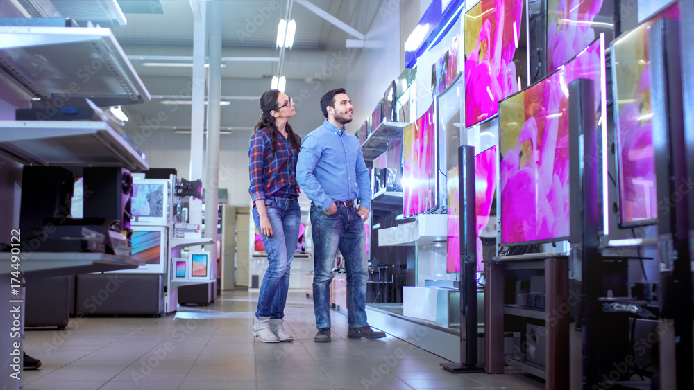 In the Electronics Store Young Couple is Choosing Which a New Model of 4K TV's is Best for Their Home and Budget. In the Electronics Store Young Couple is Choosing Which a New Model of 4K TV's is Best for Their Home and Budget.