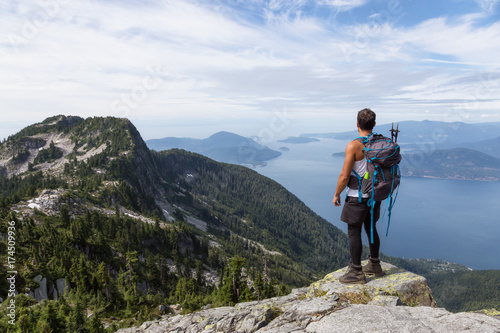 Fit and Adventurous Latin American man is hiking on top of a mountain ridge with a beautiful ocean view in the background. Taken on Lions Peaks, North of Vancouver, British Columbia, Canada. 