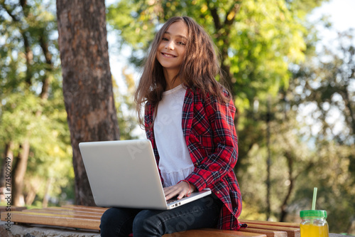 Smiling brunette young girl sitting in park with laptop computer