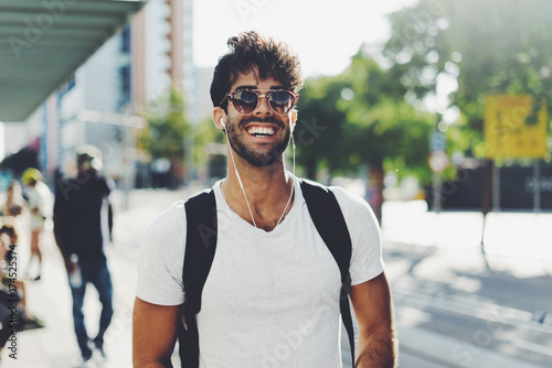 Young travel blogger is ready for a new weekend trip. Bearded backpacker is standing on a blurred station background. Student is relaxing on a weekend. Hipster guy wearing sunglasses smiling at camera photo