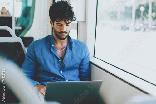 Young man is reading emails on portable computer while sitting in a city bus beside a window. Hipster guy with dark curly hair wearing light blue shirt is going to the office by a public transport.