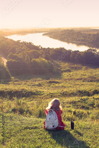 A photo of a traveler with a backpack, a girl sitting on the hill and thinking observing the sunset and the nature landscape on the hike.