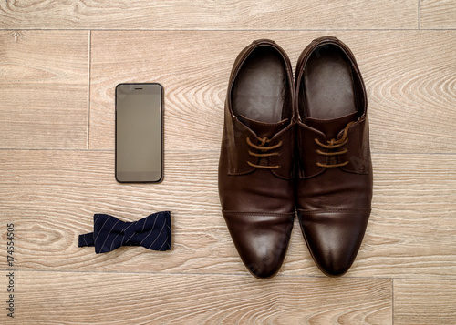 Close up of brown leather shoes men, blue bowtie and mobile phone on wood background, free space. Modern man accessories. Wedding details. Groom accessories. Men casual outfits with shoes and bowtie