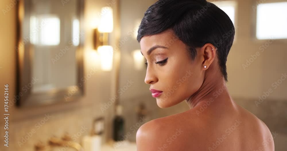 Close up of gorgeous black female with bare shoulders posing in warmly lit hotel bathroom. Portrait of lovely African American woman in her 20s looking over her shoulder 