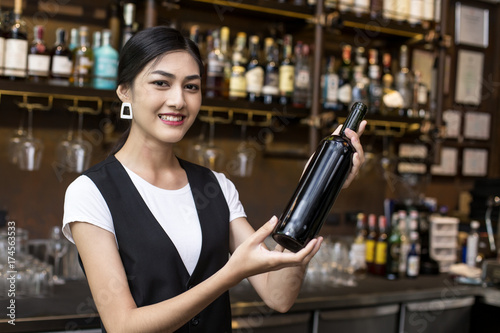 Young Asian Woman Waiter Restaurant Catering Service Happy Emotion. Woman Waiter Present Red-Wine for Customer at Bar. Woman with Wine in Bar Concept.