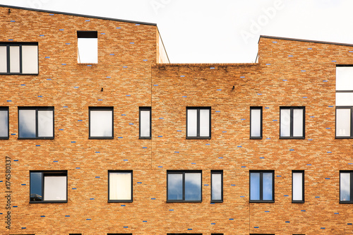 Facade of a residential orange brick building with angular rooftop, lots of windows of differente sizes with sky reflections, curtains and vases.