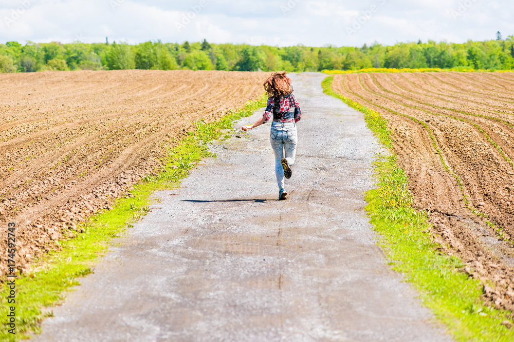 Young woman running, jumping in air and smiling on countryside dirt road by brown plowed fields with furrows in summer in Ile D'Orleans, Quebec, Canada Young woman running, jumping in air and smiling on countryside dirt road by brown plowed fields with furrows in summer in Ile D'Orleans, Quebec, Canada
