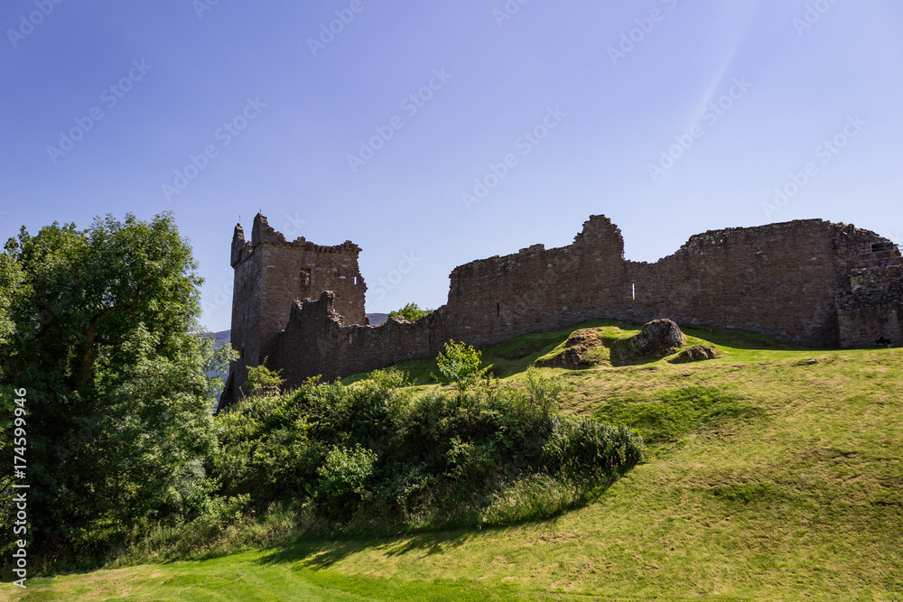 The Ruins of Urquhart Castle in the bright summer sun with sun rays and lush green foliage.