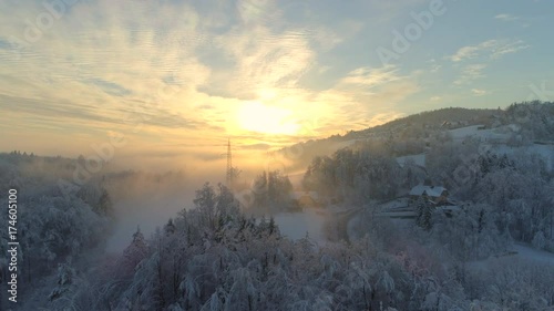AERIAL: Flying over frozen snowy treetops towards countryside town an misty sunrise. Golden sun rising behind icy mixed forest covered in morning fog and snow in cold winter. Stunning winter landscape