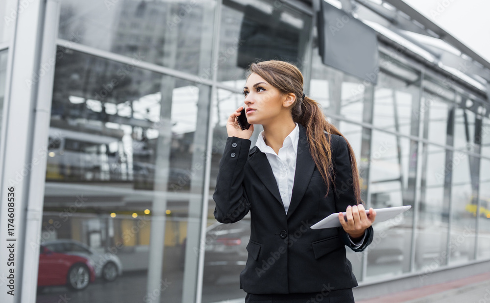 Successful businesswoman or entrepreneur using a digital tablet computer and talking on cellphone standing in front of his office.