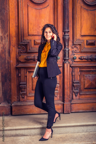 Obraz na płótnie Dressing in black blazer, orange shirt, pants, heels, a young East Indian American business woman walking down vintage style office door way, carrying laptop computer, making call on her mobile phone. w salonie