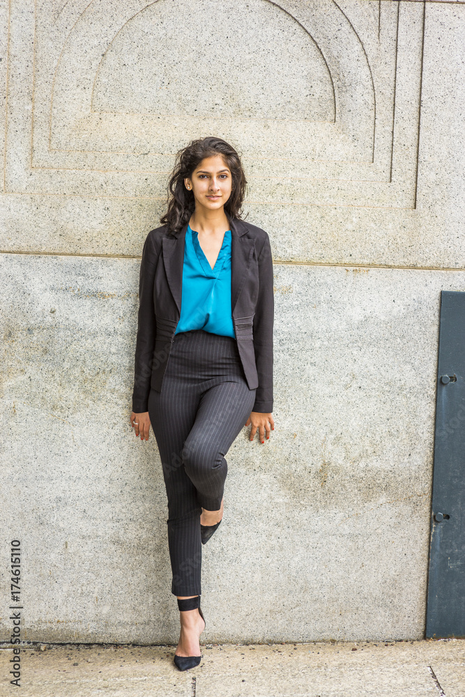 Modern East Indian American Student. Dressing in black blazer, blue under  shirt, striped pants, heels, a young girl with long curly hair standing  against wall on campus, smiling, looking at you.. Stock