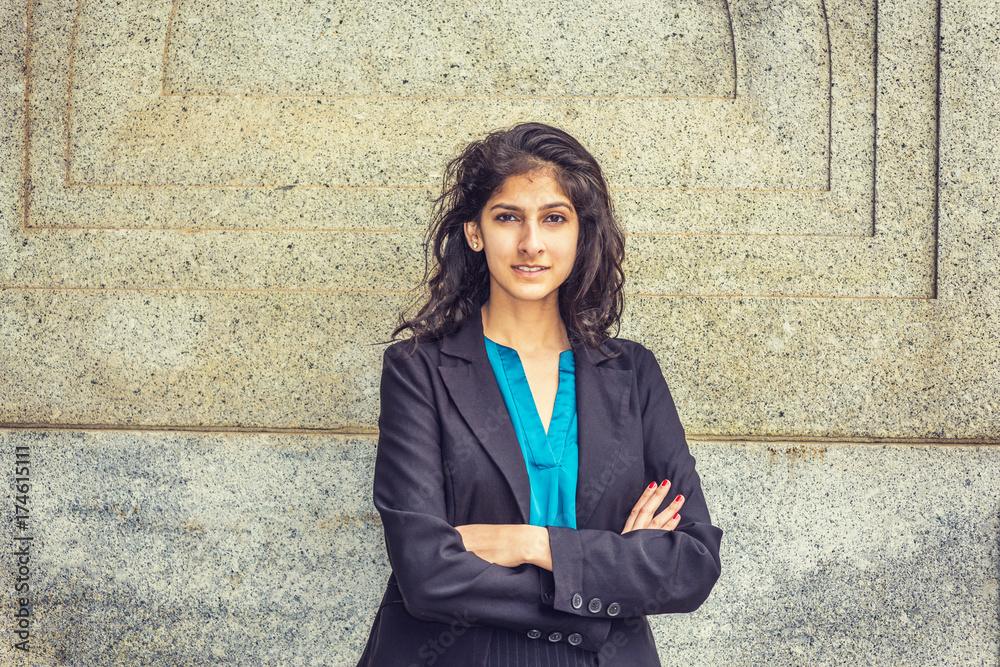 Modern East Indian American Student. Dressing in black blazer, blue under  shirt, crossing arms, a young girl with long curly hair standing by wall,  smiling, looking at you. Instagram filtered effect. Stock